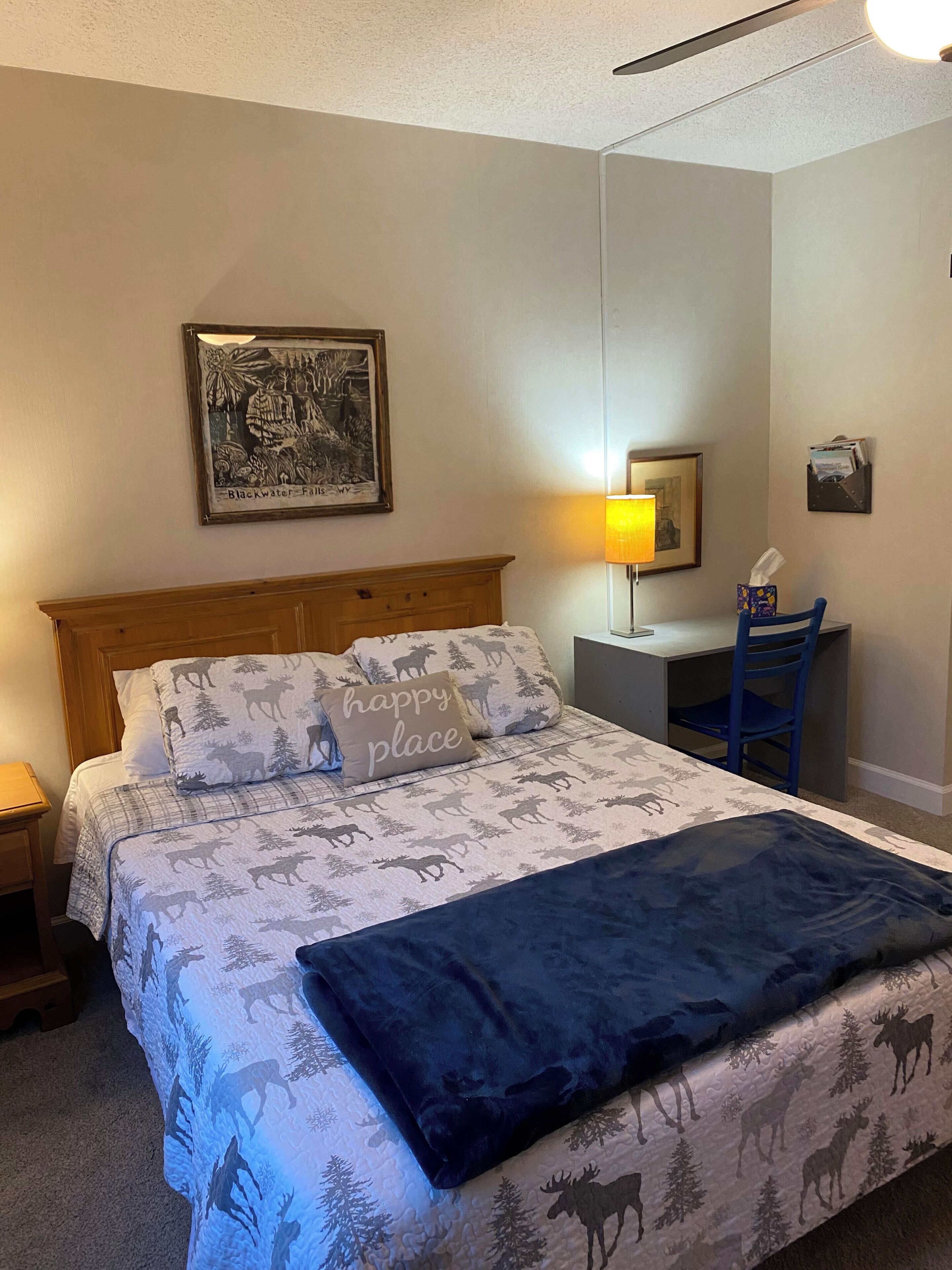Une chambre à coucher avec un lit recouvert d’une courtepointe à motifs d’élans, une tête de lit, un bureau avec une chaise, une lampe et un tableau encadré accroché au mur.