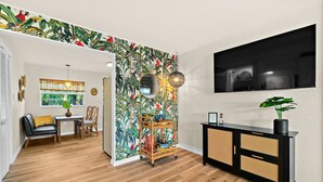 A room with a wooden floor, a black console table with a plant, a wall with a tropical wallpaper, and a large TV mounted on the wall. - Boutique style Apt/Walk to Beach/Pet friendly (Indian Harbour Beach)