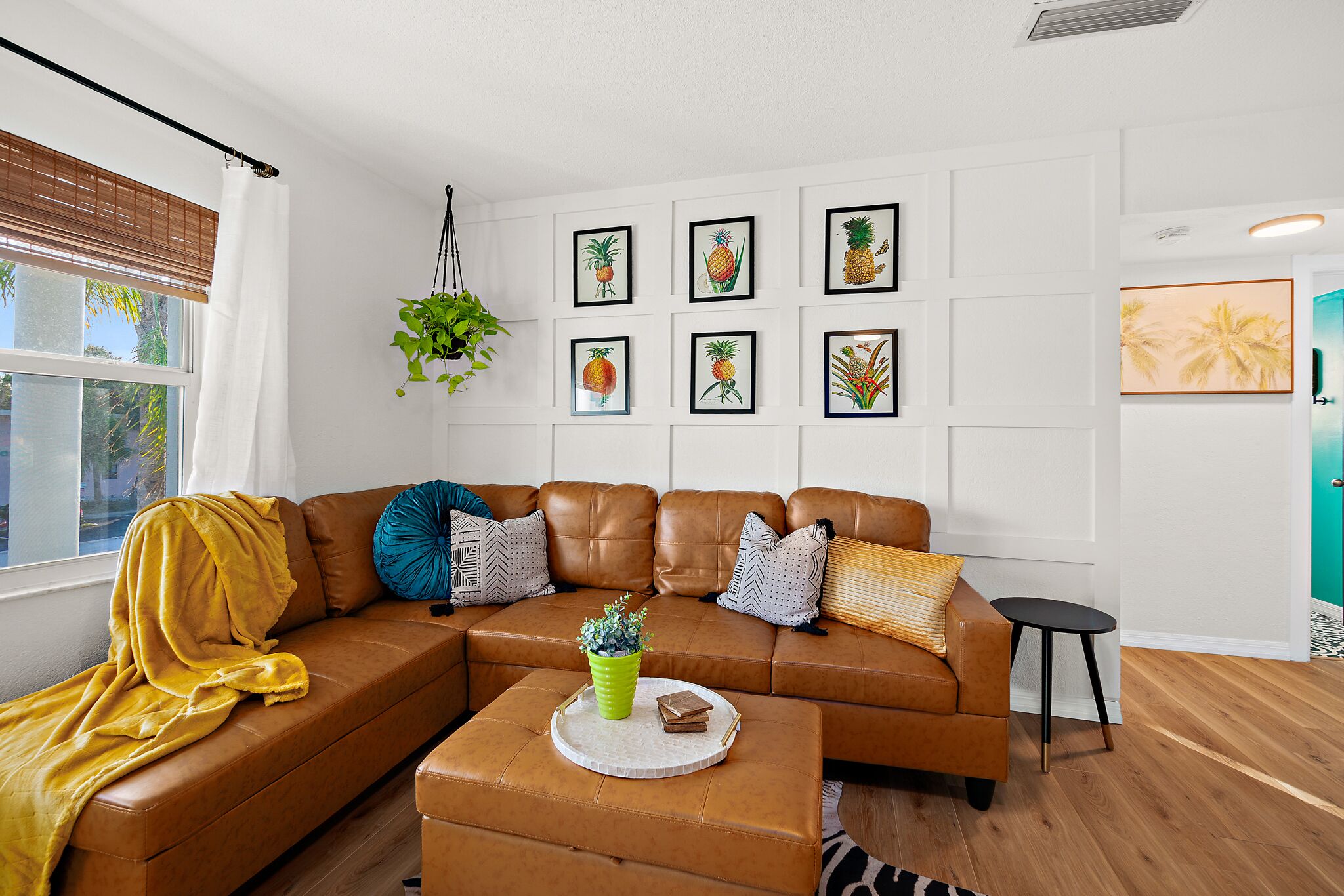 Una sala de estar moderna con un sofá de cuero marrón, una manta amarilla, una planta verde y un cuadro con frutas tropicales en la pared.