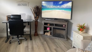 A living room with a TV displaying a beach scene, a desk with a chair, a wooden vase with dried plants, and a small cabinet with decorative items. - Classy, Comfy and Cozy (Timmins)
