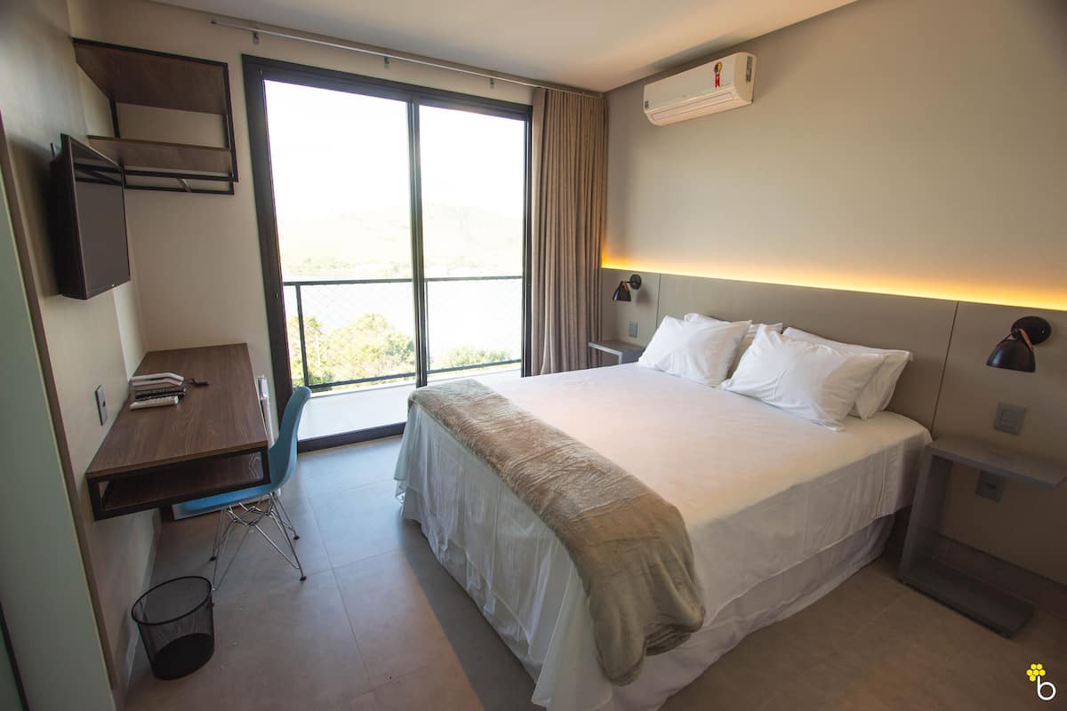 A hotel room with a large bed, a desk, and a chair. There is a window with curtains, an air conditioning unit on the wall, and a trash can.