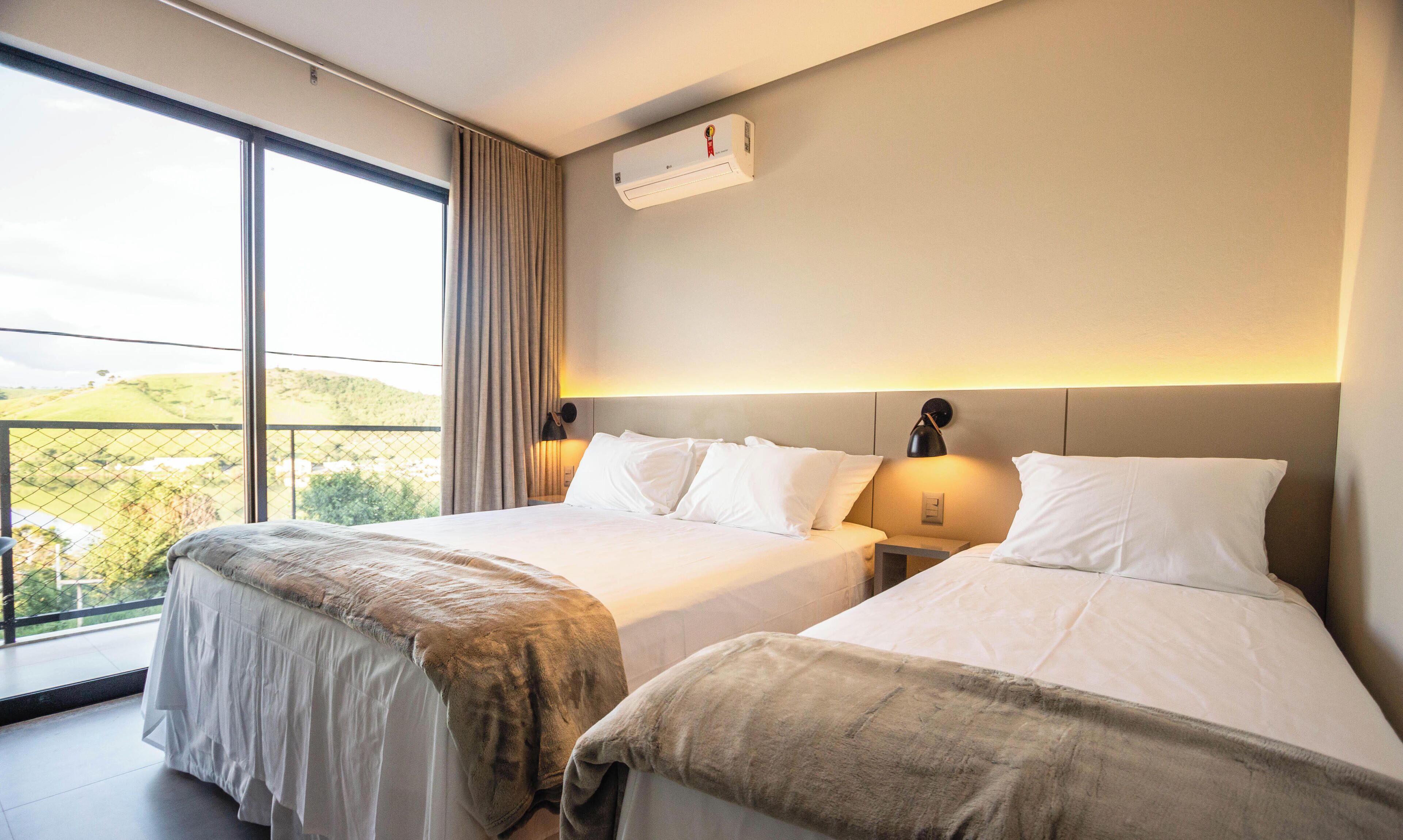 Habitación de hotel con dos camas, un ventanal con vistas a zonas verdes, un equipo de aire acondicionado y una mesita de noche con lámpara.
