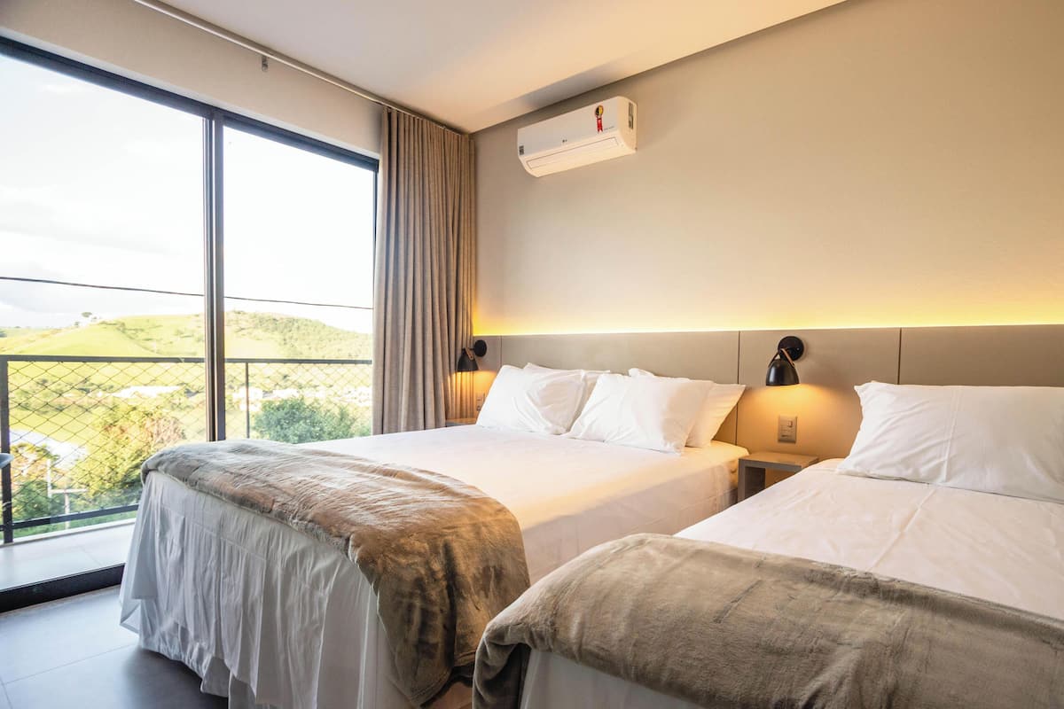 A hotel room with two beds, a large window with a view of greenery, an air conditioning unit, and a nightstand with a lamp.
