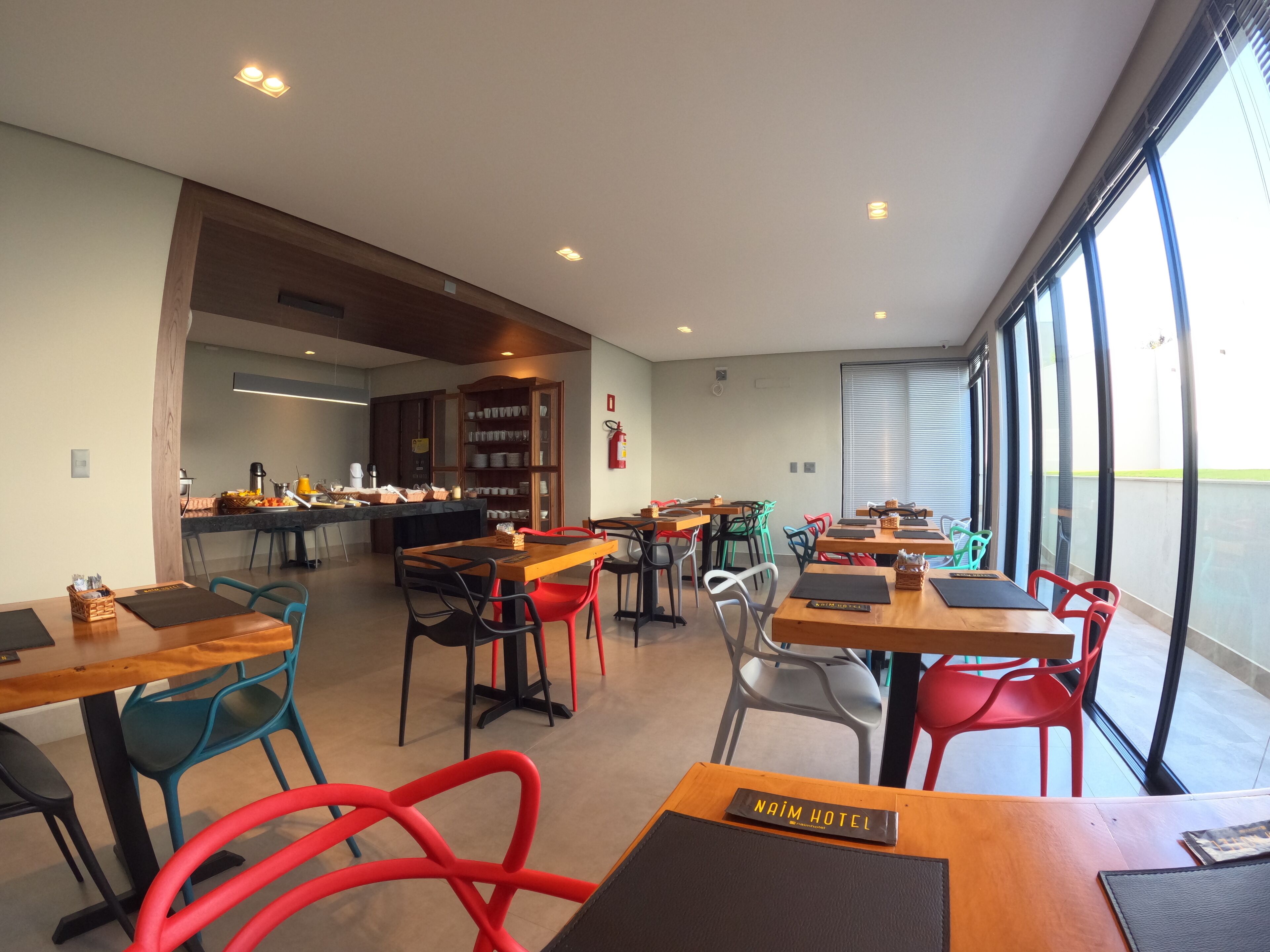A modern hotel lobby with wooden tables and chairs, a kitchen area, and large windows offering views of the outdoors.