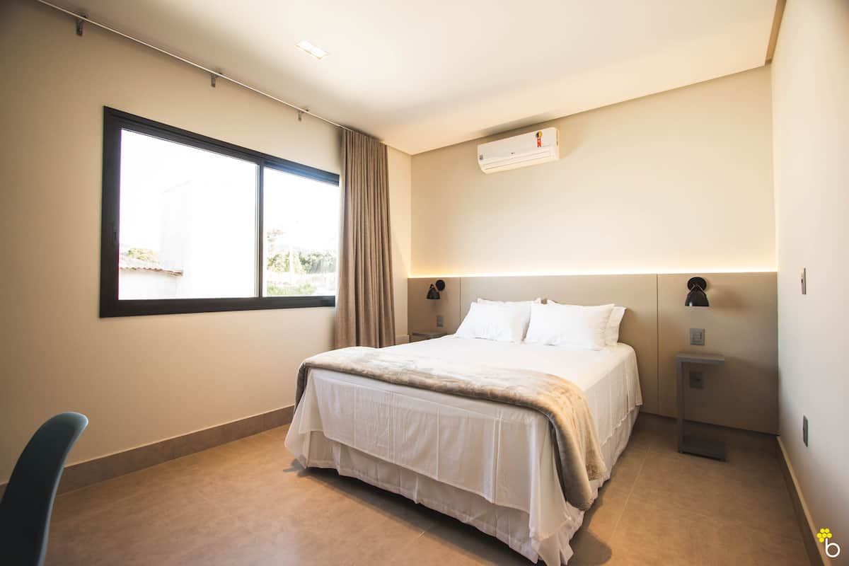 A bedroom with a large bed, a window, an air conditioner, and a bedside table with a lamp.