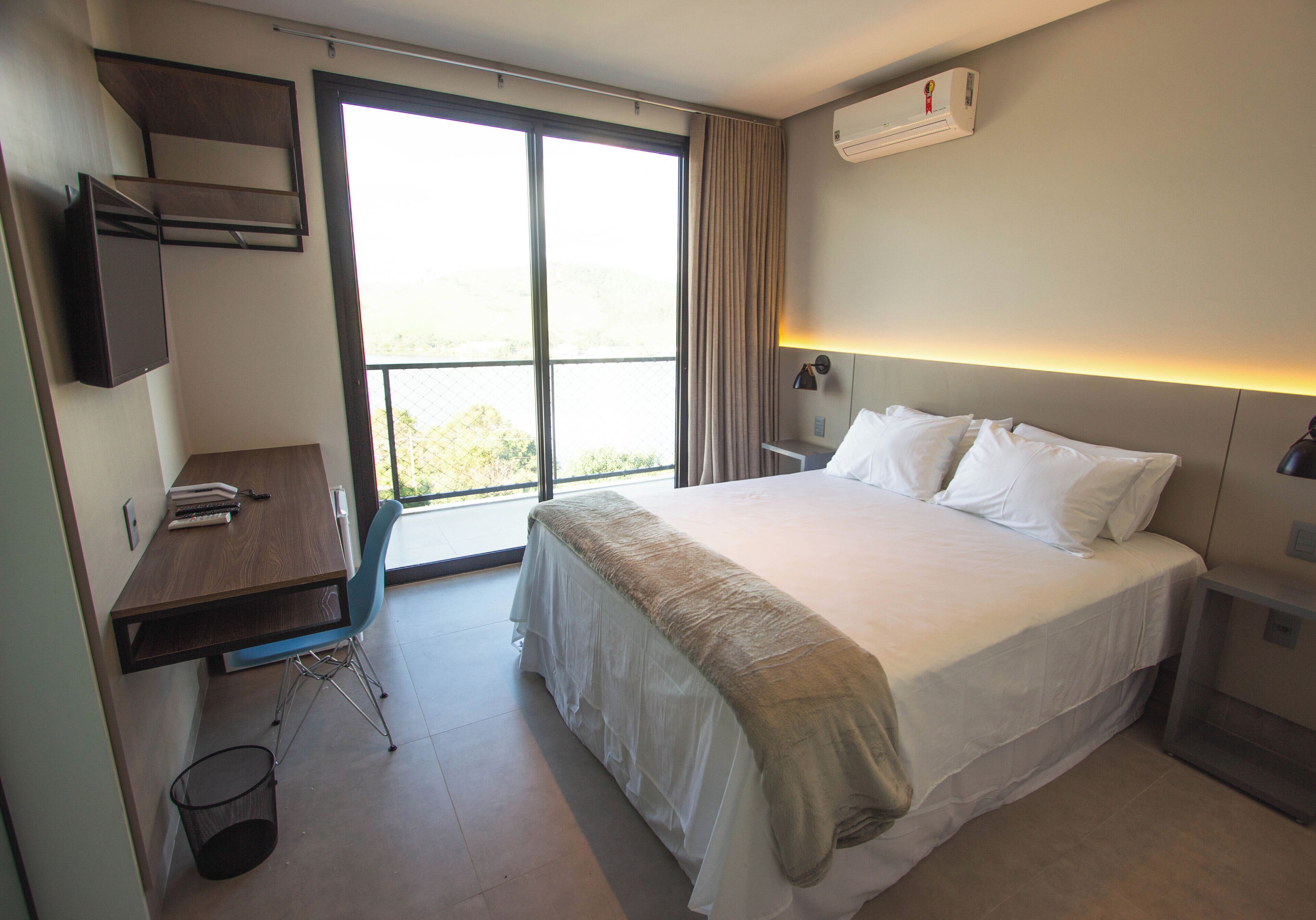 Habitación de hotel con una cama grande, un escritorio y una silla. Hay una ventana con cortinas, un equipo de aire acondicionado en la pared y un bote de basura.
