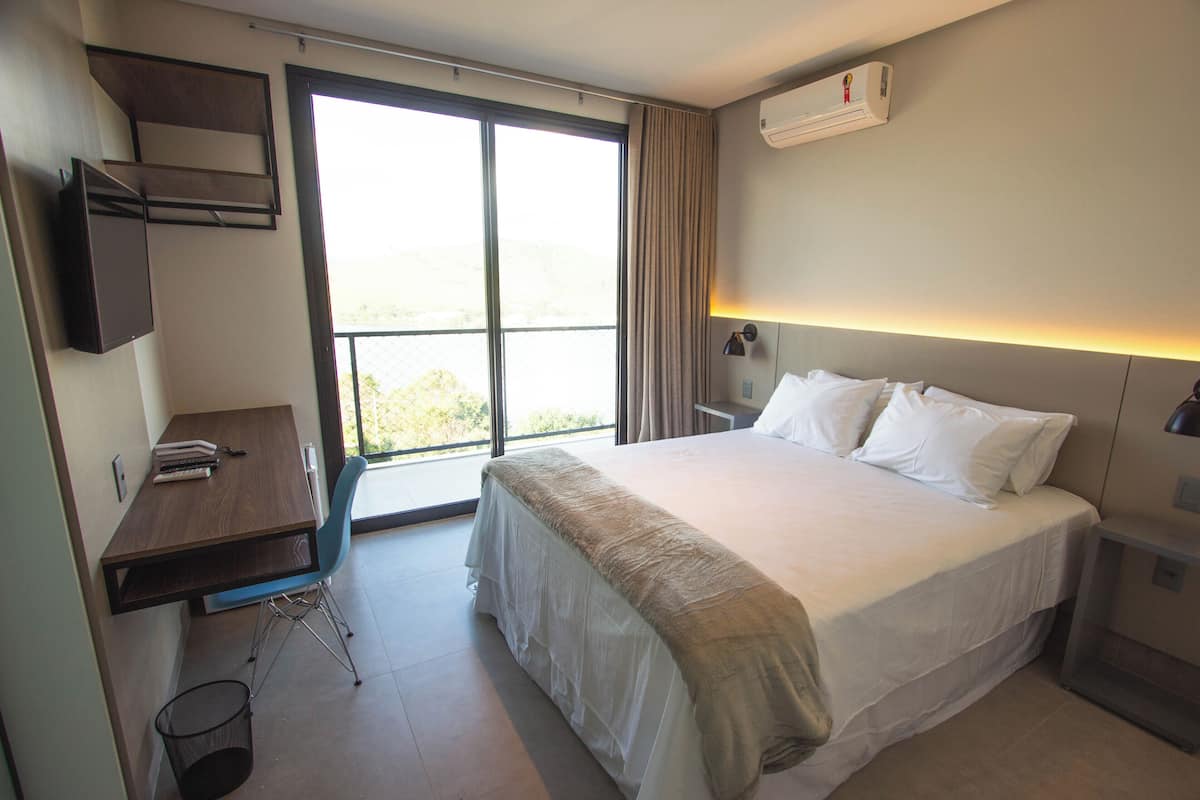 A hotel room with a large bed, a desk, and a chair. There is a window with curtains, an air conditioning unit on the wall, and a trash can.
