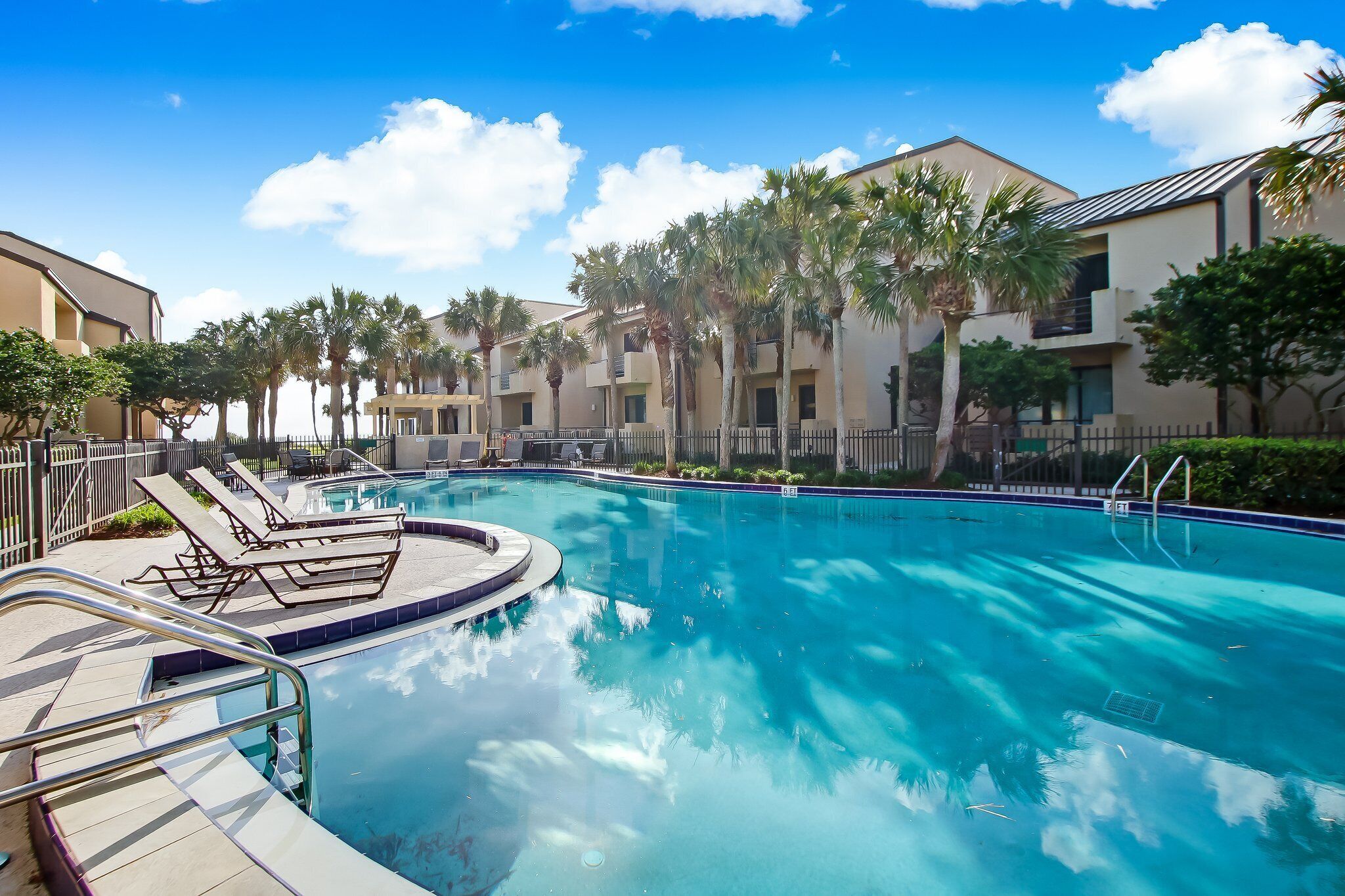 A swimming pool with lounge chairs and a poolside area, surrounded by palm trees and buildings.