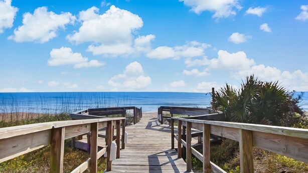 Une promenade en bois qui mène à une plage, avec un ciel bleu clair et des nuages blancs.