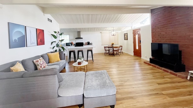 Een moderne woonkamer met een grijze hoekbank, een salontafel en een televisie tegen een bakstenen muur. Op de achtergrond is een keuken met een bargedeelte en barkrukken zichtbaar.