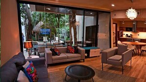 Ein modernes Wohnzimmer mit einer Couch, Sesseln und einem Couchtisch. Es gibt einen Essbereich mit Tisch und Stühlen. Der Raum verfügt über große Fenster und einen Blick ins Freie.