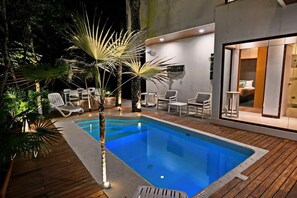 Une piscine avec des chaises longues, une terrasse en bois et un intérieur moderne, avec un lit visible à travers une porte vitrée.