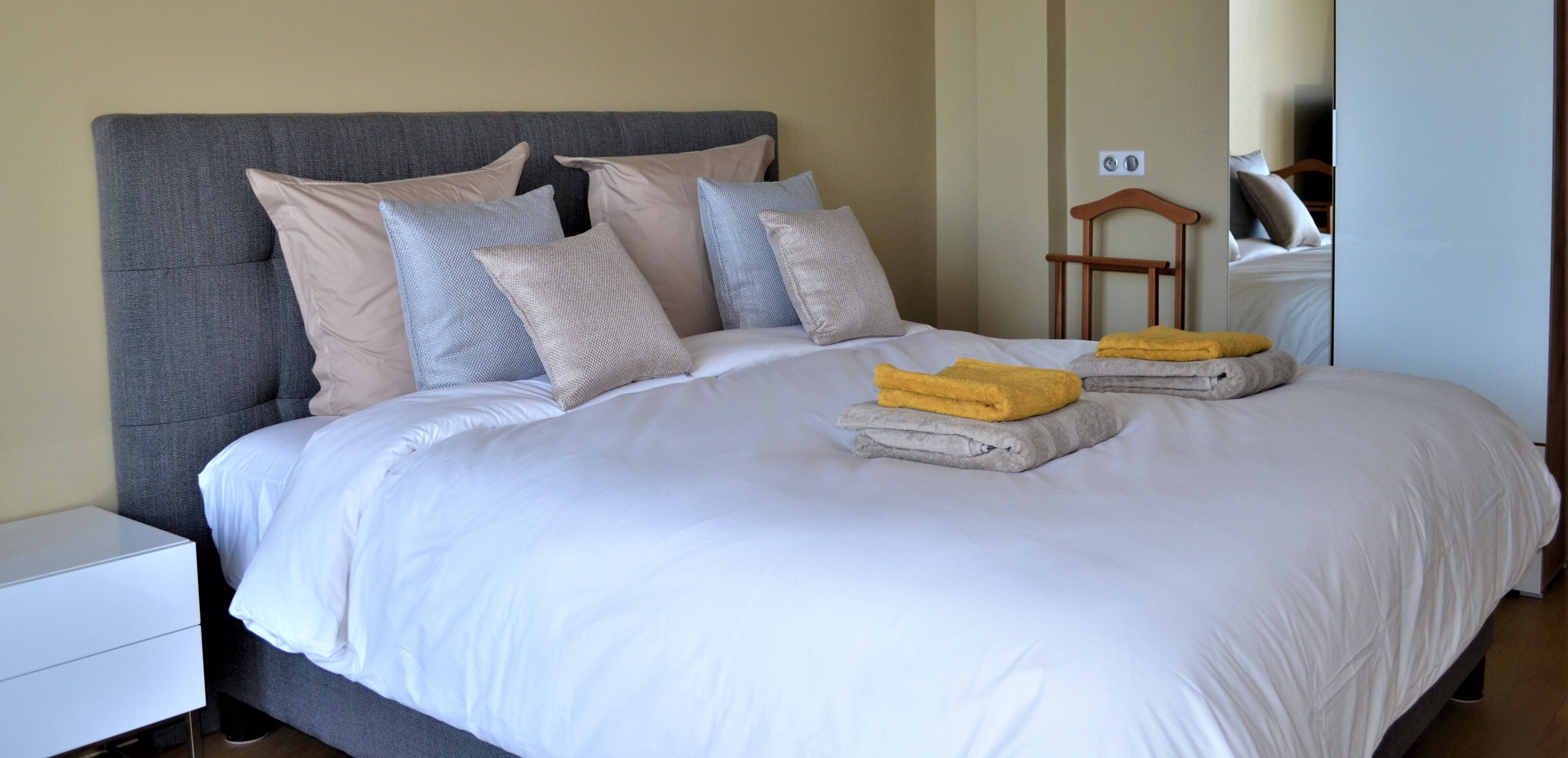 A neatly made bed with white linens and multiple pillows, a wooden nightstand, and a mirror reflecting part of the room.