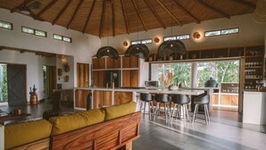 A spacious kitchen with a wooden bench, bar stools, and a large window overlooking greenery. - La Musa de la Jungla (Nosara)