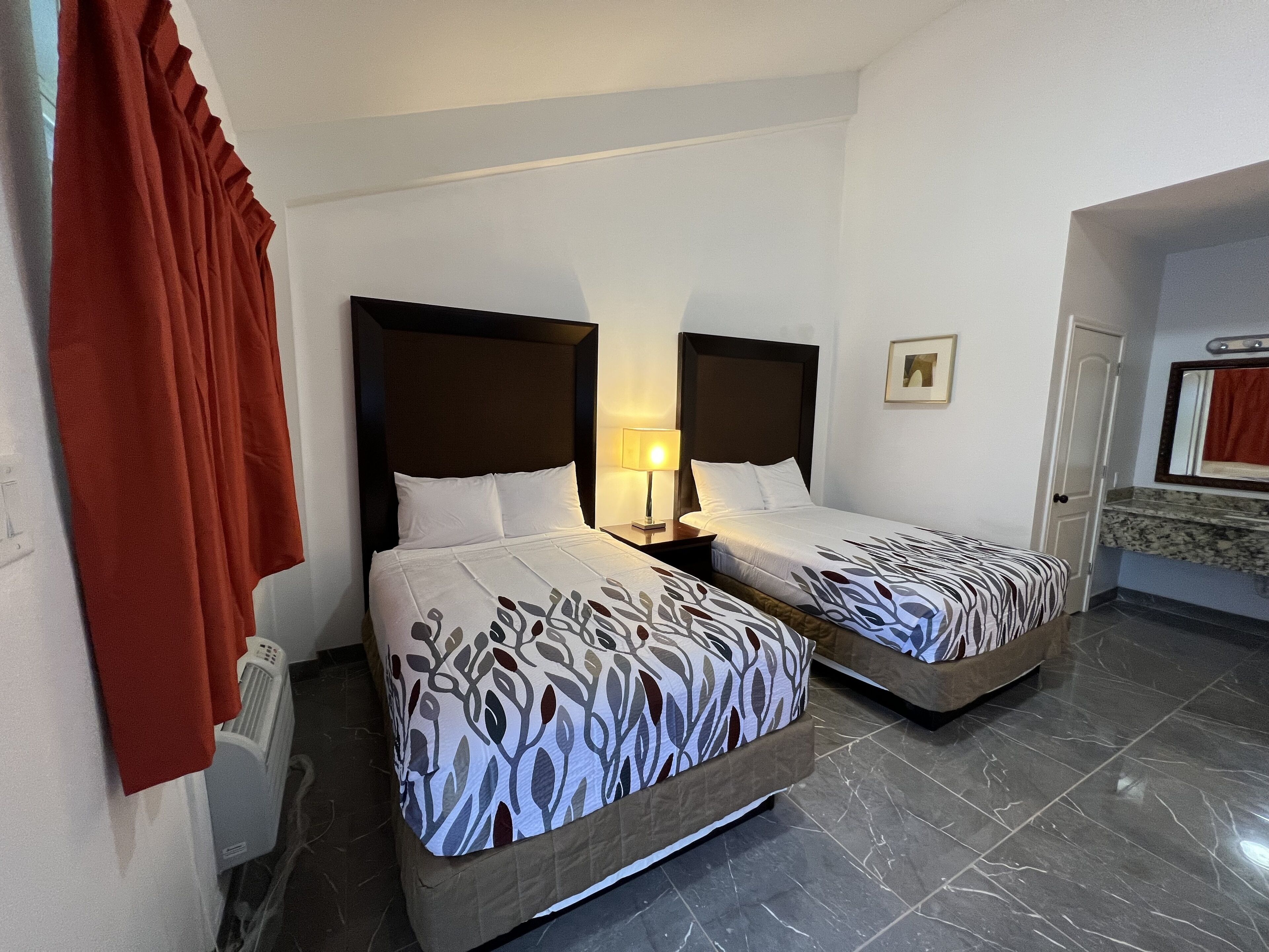 Een hotelkamer met twee bedden, een nachtkastje met een lamp, een badkamer met een spiegel en een rood gordijn.