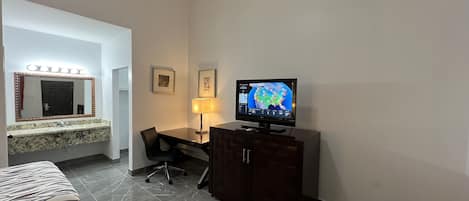 A hotel room with a bed, a desk with a chair, a television, a mirror, and a cabinet.
