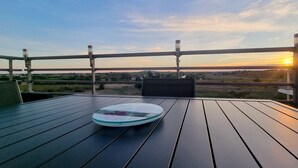 Um deck de madeira com mesa e cadeiras de vidro, com vista para uma paisagem com pôr do sol.