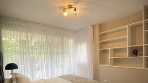 Un dormitorio con cama, lámpara de noche, ventana con cortinas translúcidas y estanterías empotradas.