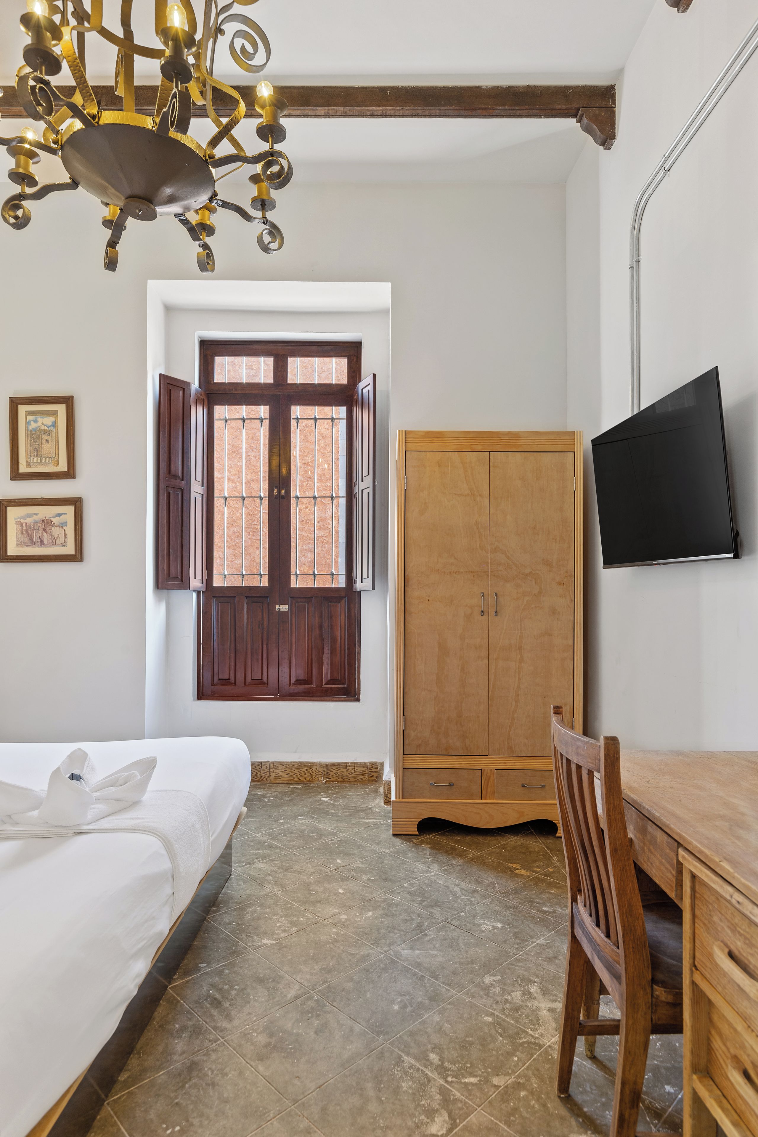 A bedroom with a bed, a wooden wardrobe, a television mounted on the wall, and a window with shutters.