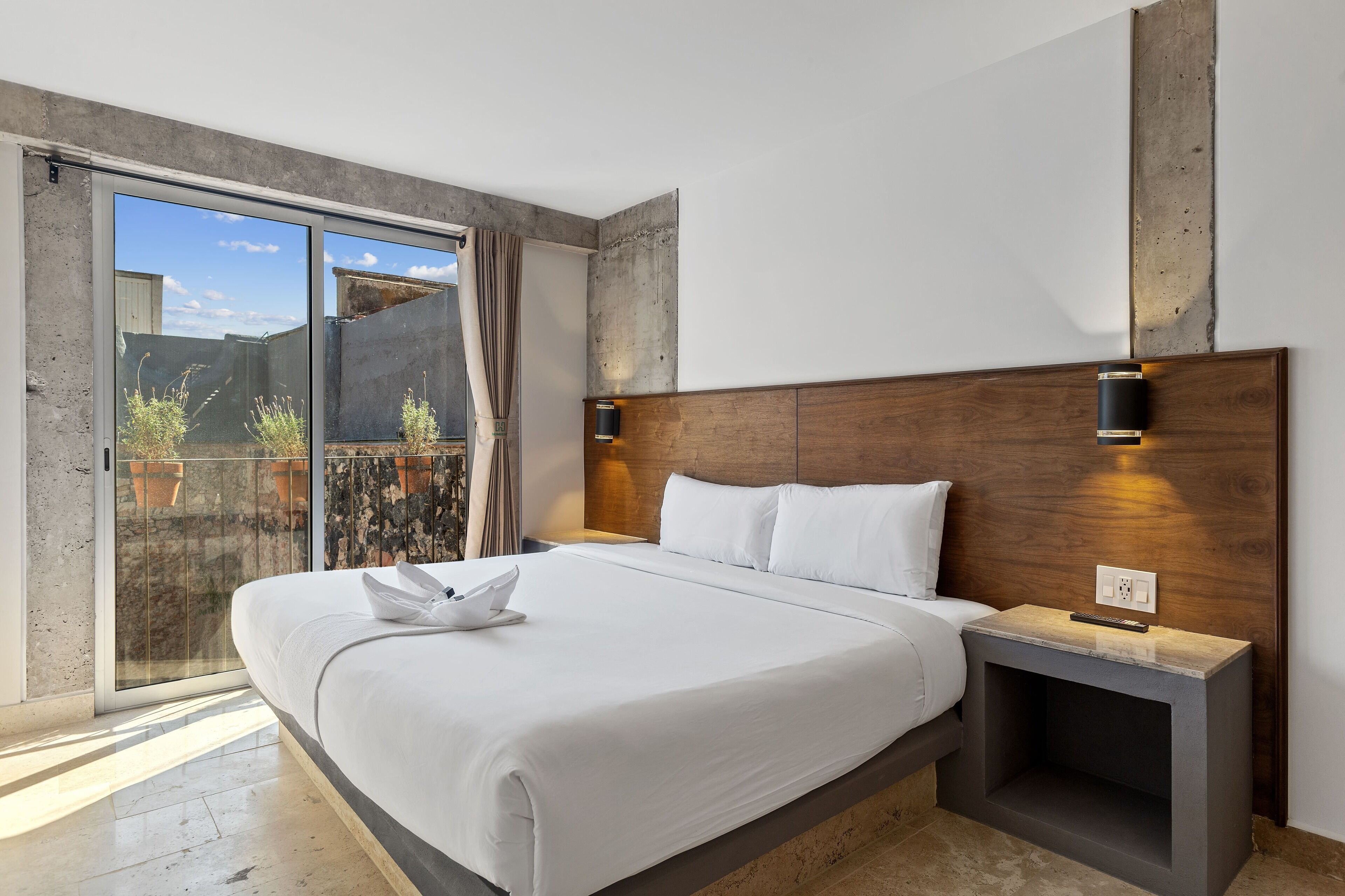 A modern hotel room with a large bed, a wooden headboard, a nightstand, and a sliding glass door leading to an outdoor area.