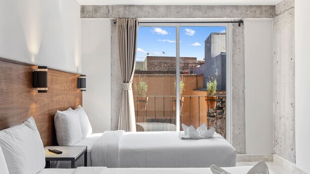 Ein Hotelzimmer mit zwei Betten, einem Holzkopfende, einem Nachttisch, einem Fenster mit Vorhängen und Blick auf die Stadtlandschaft.
