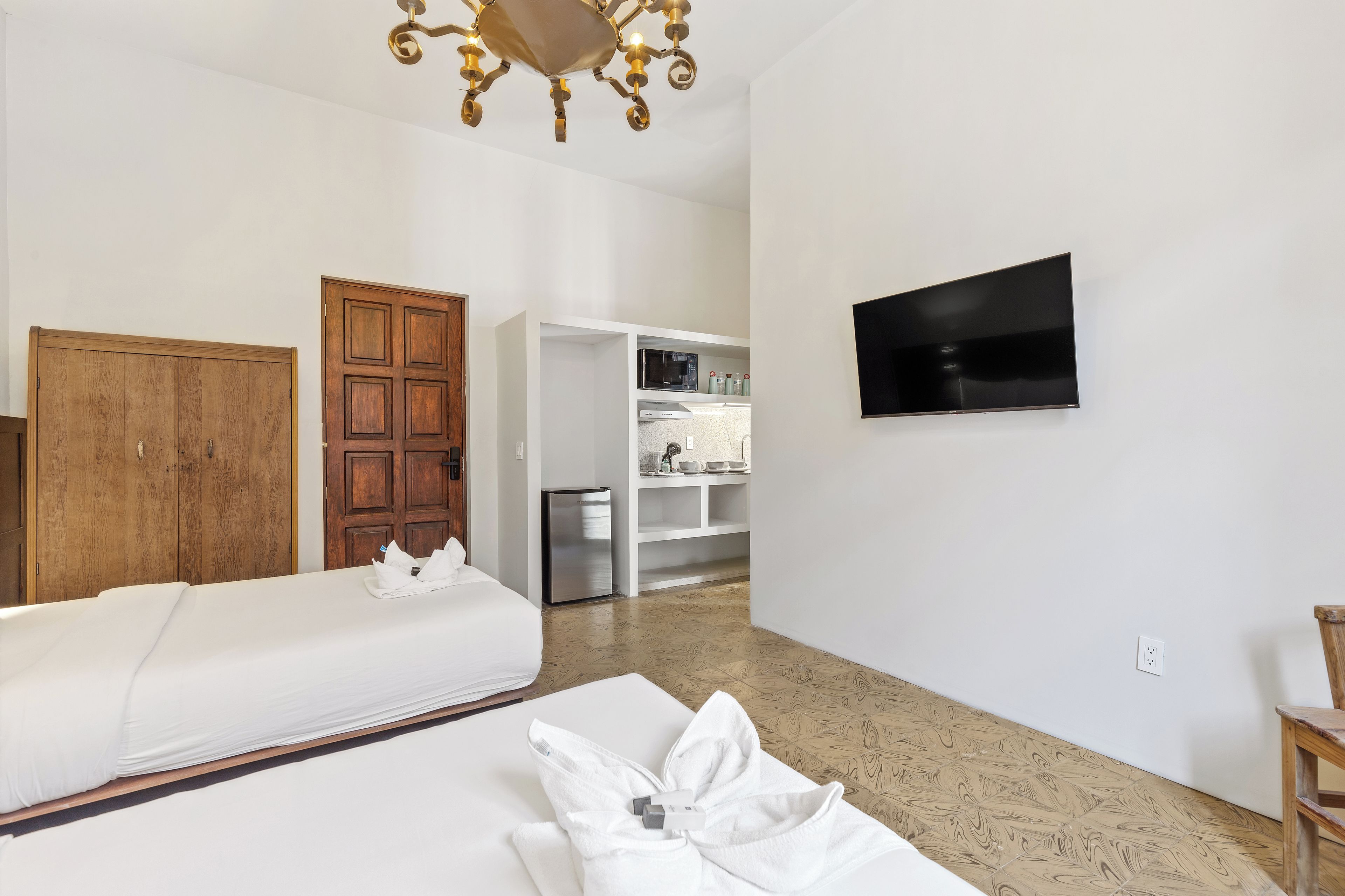 A hotel room with two beds, a wooden wardrobe, a kitchenette, and a flat-screen TV.