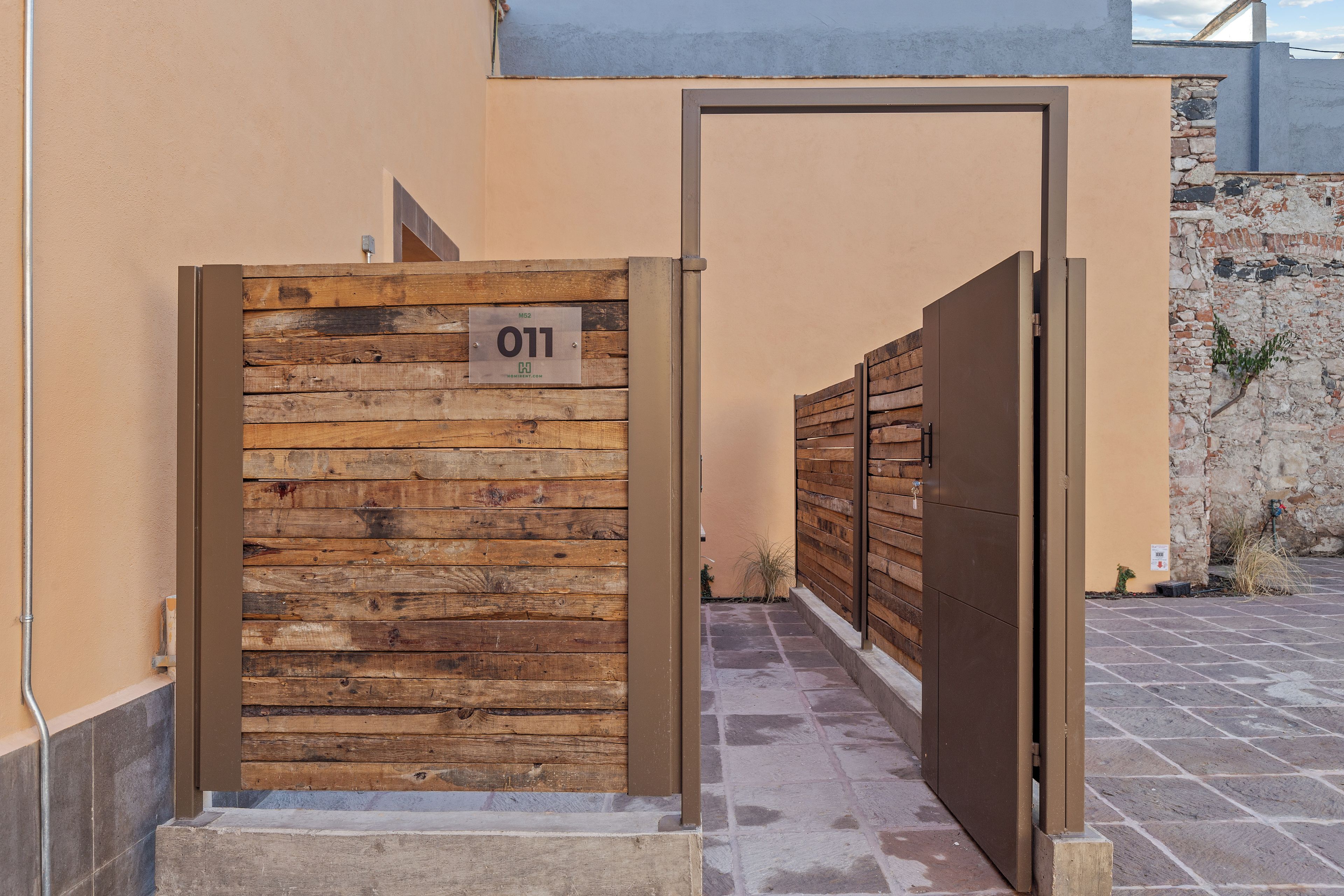 A gated entrance with a wooden panel and a metal gate, a sign with the number '011', and a stone wall in the background.