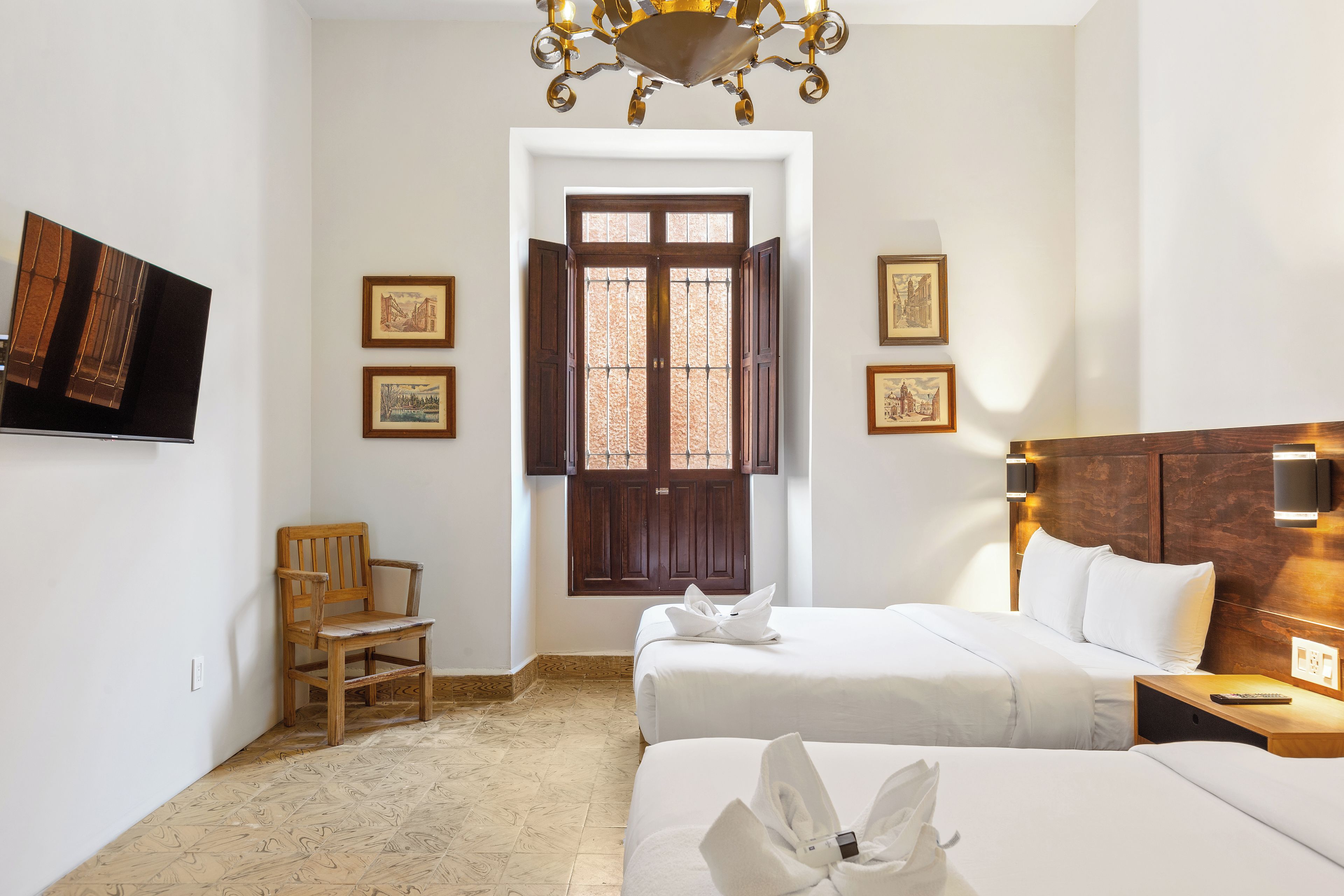 A hotel room with two beds, a wooden chair, a desk with a lamp, and a large window with shutters.