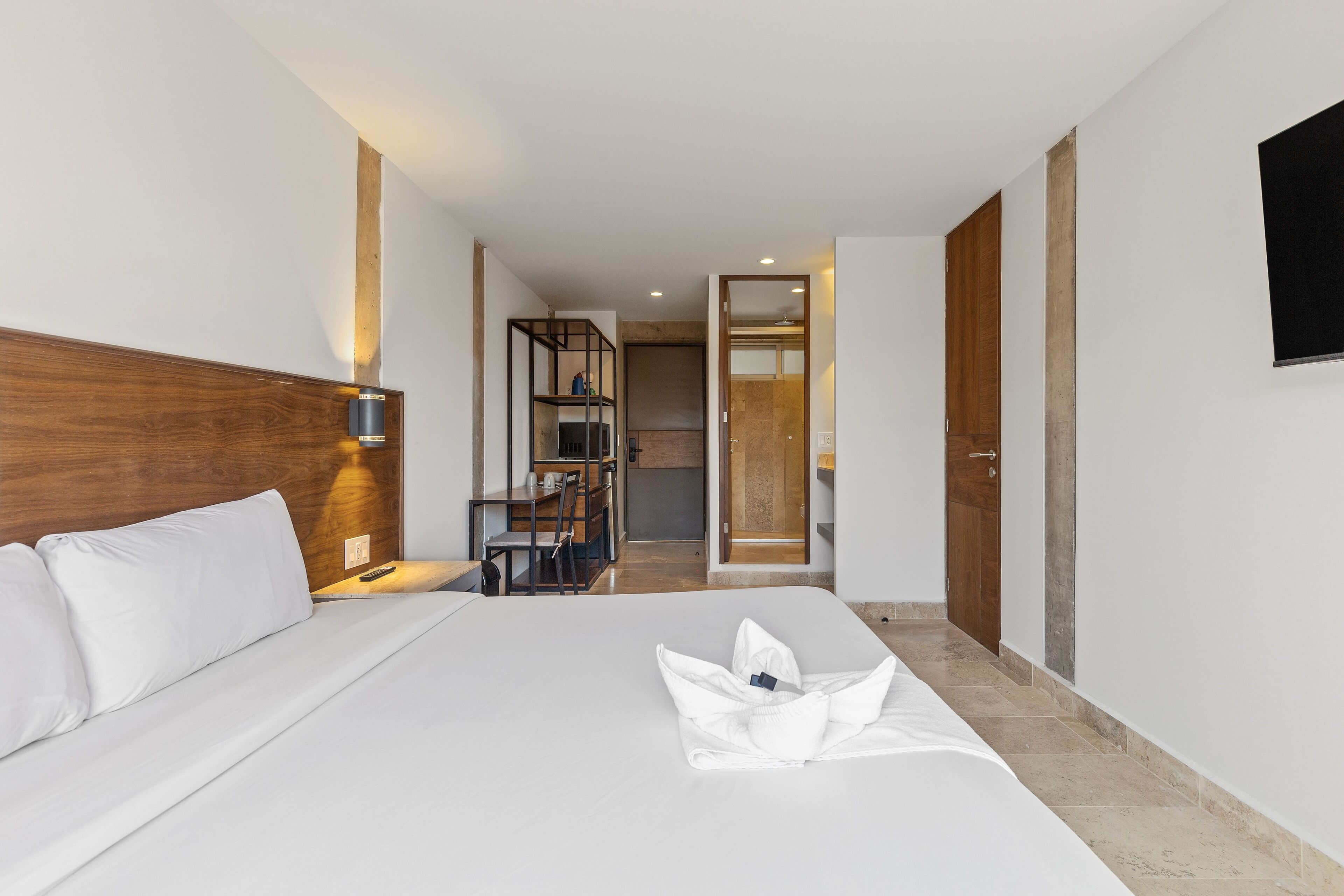 A hotel room with a large bed, a wooden headboard, a bedside table with a lamp, a wardrobe, and a television mounted on the wall.