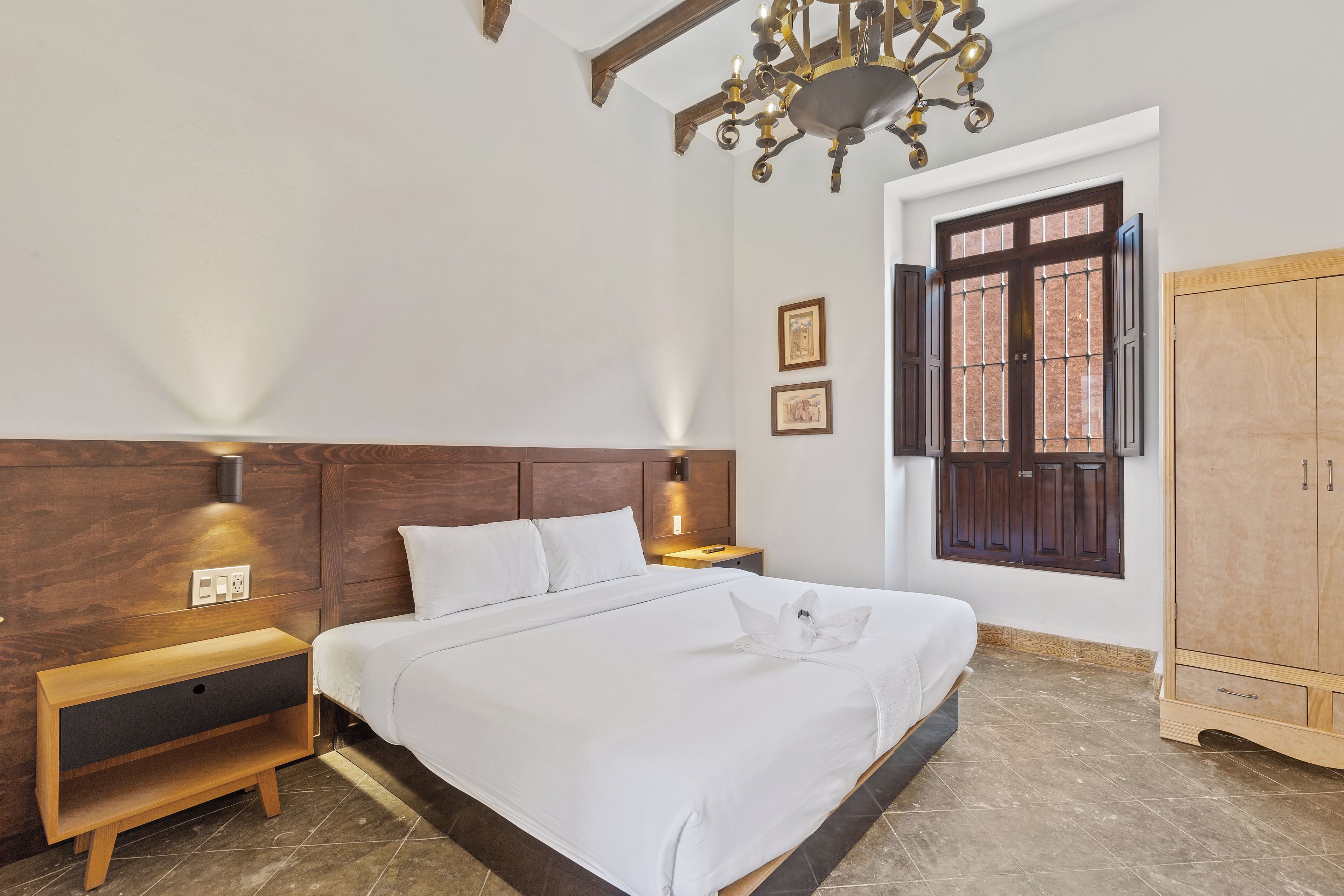 A hotel room with a bed, bedside table, nightstand, wall-mounted light, wooden wardrobe, and a window with a view.
