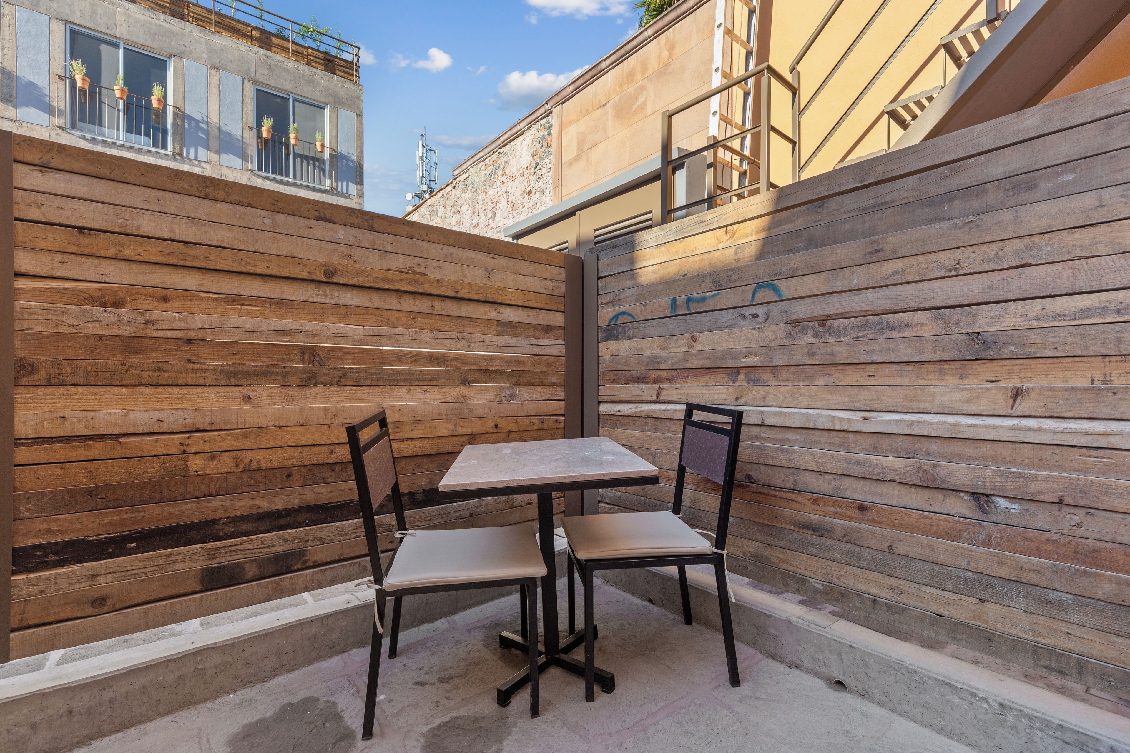 An outdoor seating area with a wooden table and chairs, a concrete floor, and a view of a building with a stone facade.