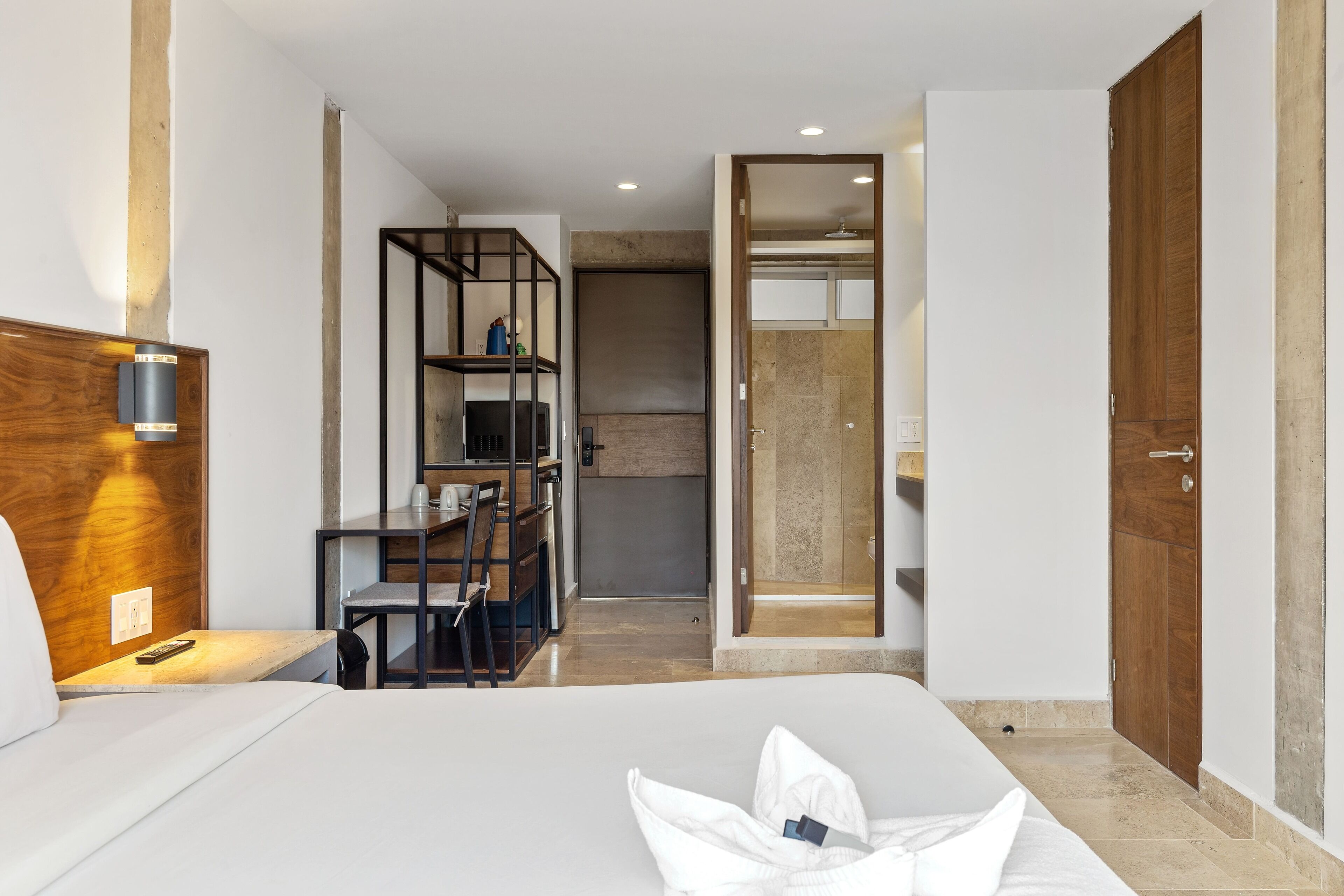 A modern hotel room with a bed, a desk, a chair, a wardrobe, and a bathroom visible through an open door.