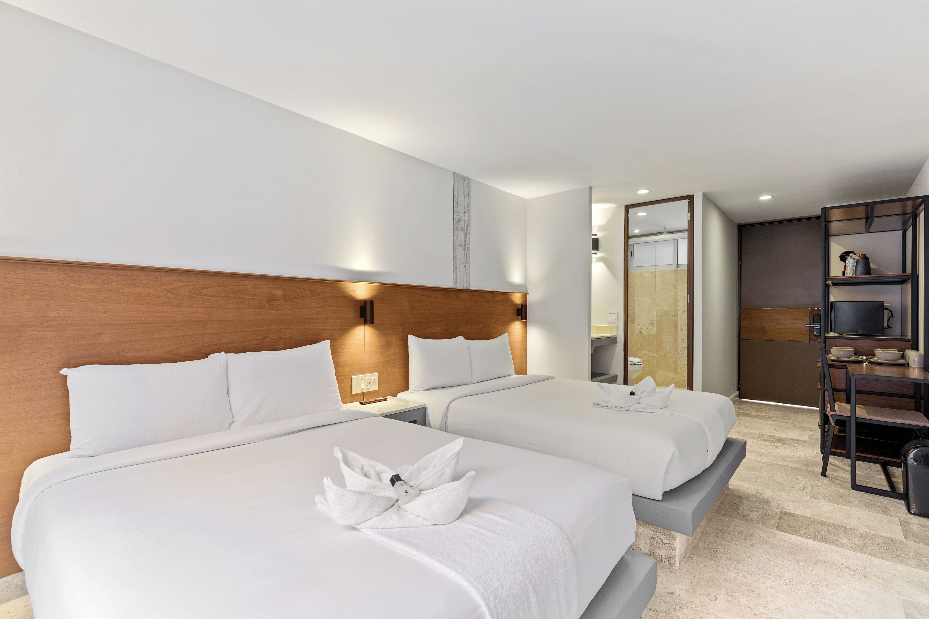 A hotel room with two beds, a wooden headboard, a small table with a lamp, and a kitchenette area.