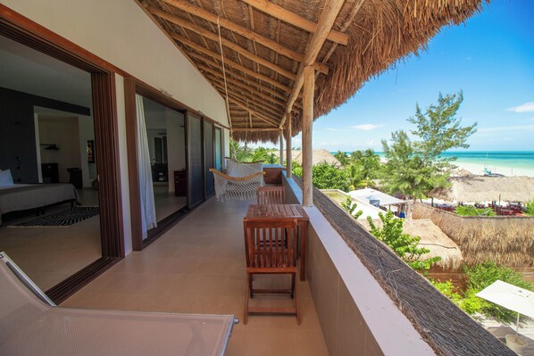 A balcony with a view of the beach, wooden furniture, and a thatched roof.