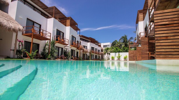 A pool area with clear water, wooden deck, and white buildings with balconies.