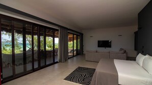 A modern bedroom with a large bed, a sofa, and a TV. It features a sliding glass door leading to an outdoor area with a table and chairs.