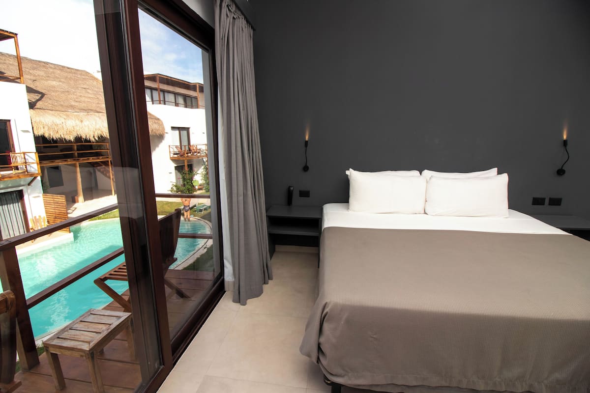 Quarto de hotel com uma cama grande, vista para uma piscina e uma varanda com telhado de palha.