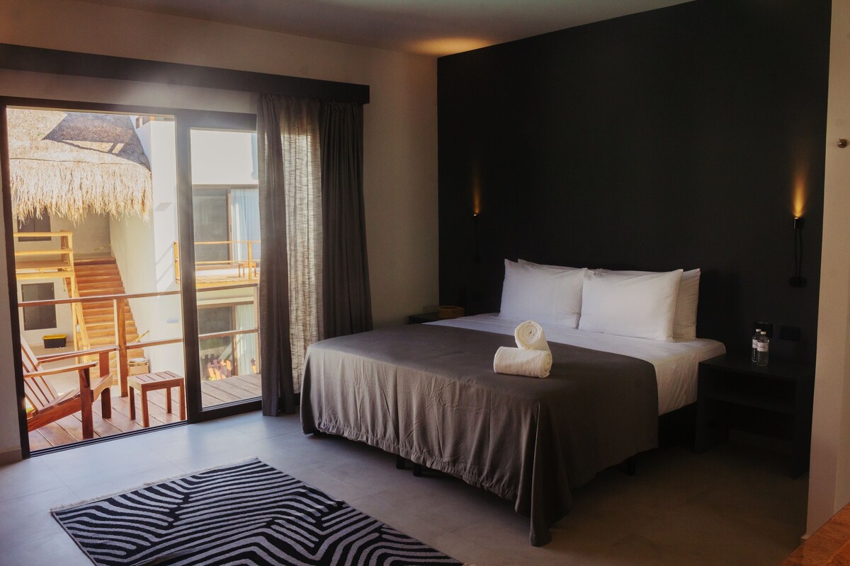 A bedroom with a bed, a nightstand, a lamp, a window with curtains, and a view of a deck area.