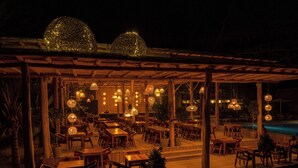 An outdoor dining area with wooden tables and chairs, hanging wicker lamps, and a pool in the background.