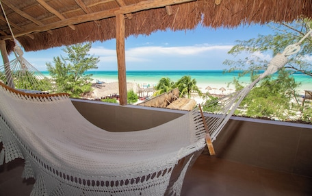 A hammock under a thatched roof overlooking a beach with palm trees and a clear sky.