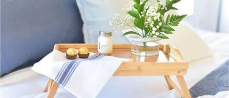 Ett brickbord med en burk grädde, en muffin och en vas med blommor, placerat på en säng med ett vitt och blått täcke.