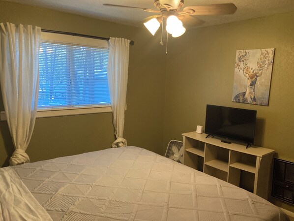 A bedroom with a bed, a ceiling fan, a TV on a stand, a window with curtains, and a painting on the wall.