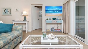 Een woonkamer met een bank, een salontafel met decoratieve objecten, een televisie waarop een strandscène te zien is, en een schuifglazen deur die leidt naar een balkon.
