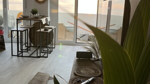 A modern living room with a large window overlooking a balcony, a dining area with a table and chairs, and a kitchen area visible through the window. - BALERMA BEACH (El Ejido)