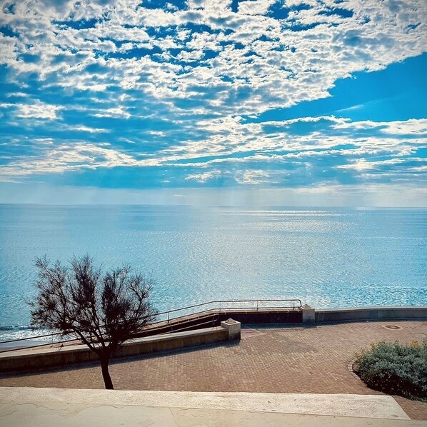 Vista al mar con cielo despejado, mar tranquilo y zona pavimentada con un árbol y un pequeño puente.