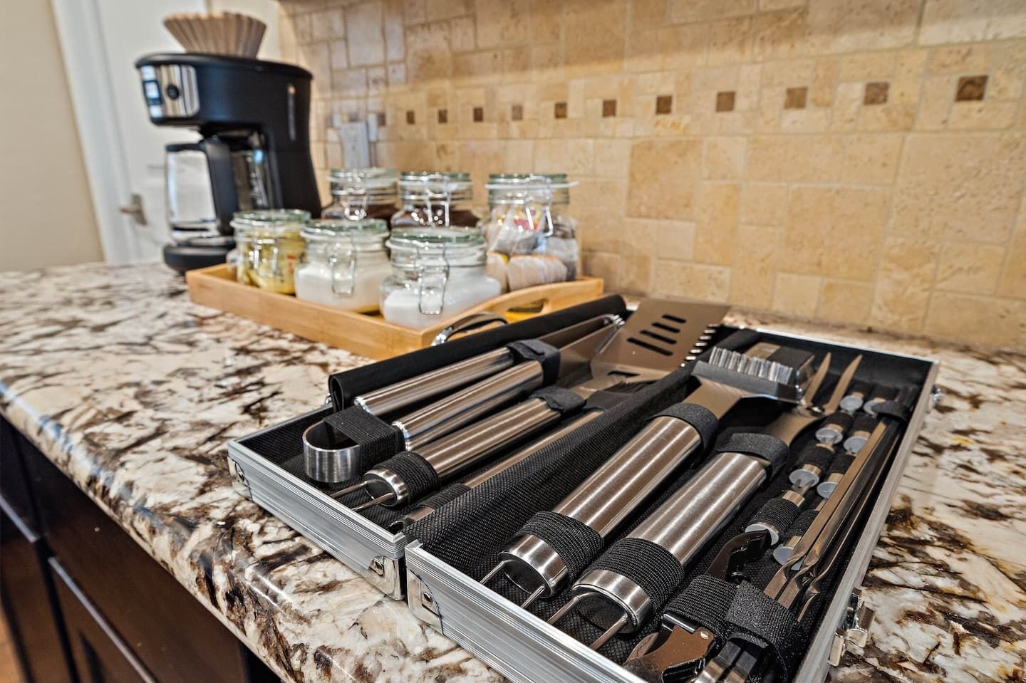 Un plan de travail de cuisine avec un ensemble d’ustensiles de cuisine en acier inoxydable rangés dans un étui, une machine à café et des pots d’épices.