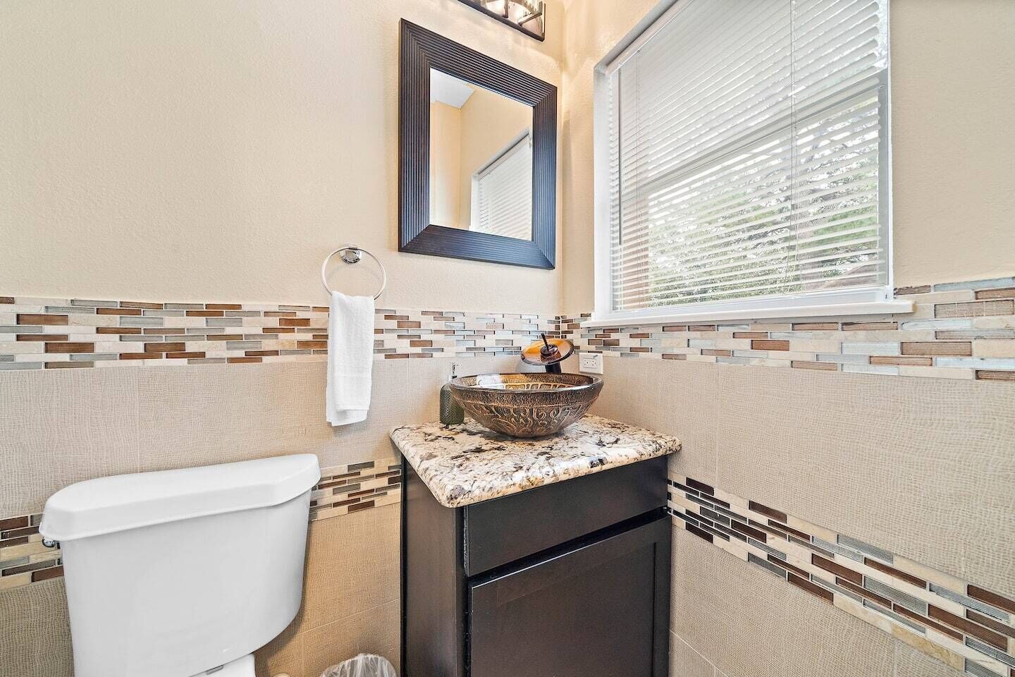A bathroom with a toilet, a sink with a decorative bowl, a mirror, and a window with blinds.