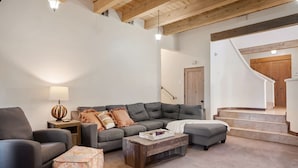 Un salon moderne avec un canapé gris, une table basse en bois et un pouf à motifs. On aperçoit à l’arrière-plan un escalier avec des marches en bois et une porte en bois.
