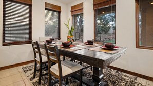 Un coin repas avec une table en bois, des chaises et une vue sur l’extérieur grâce à de grandes fenêtres équipées de stores.
