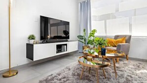 A modern living room with a flat-screen TV, white cabinetry, a round glass coffee table, a fluffy rug, and a large window with curtains.